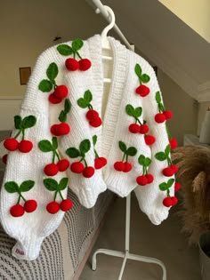 a white sweater with cherries on it hanging from a clothes rack next to a bed