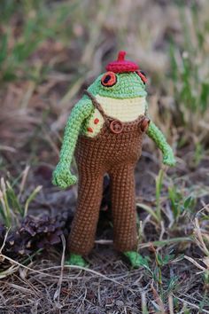 a small crocheted frog with a red hat and brown pants standing in the grass