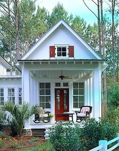 a small white house with red doors and shutters on the front porch, surrounded by greenery