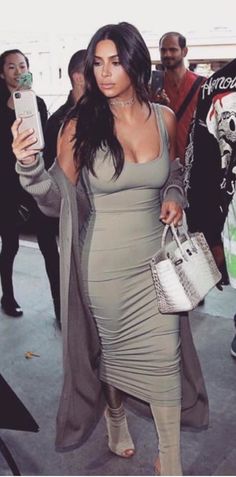 a woman in a gray dress is holding a white purse and looking at her cell phone