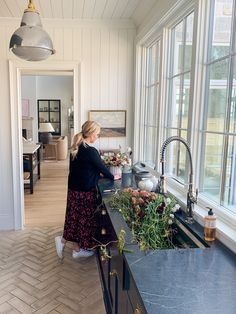 a woman standing at the sink in a kitchen next to a window with lots of windows