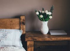 a vase with white flowers sitting on a small wooden table next to a bed in a bedroom