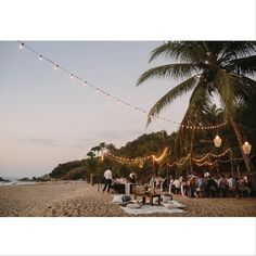 a group of people standing on top of a sandy beach next to the ocean with lights strung over them