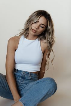 a beautiful young woman sitting on the ground wearing jeans and a white tank top with her hands in her pockets