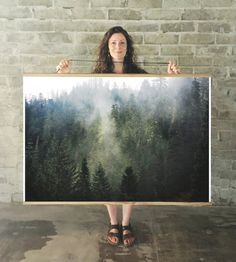 a woman holding up a large painting in front of her face with trees on it