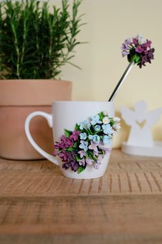 a white cup with purple and blue flowers in it on a table next to a potted plant