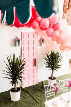 a table with pink and green balloons hanging from the ceiling next to potted plants