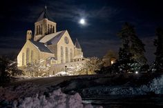 an old church lit up at night with the moon in the sky above it and snow on the ground