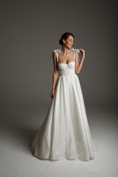 a woman in a white wedding dress standing against a gray background with her hand on her shoulder