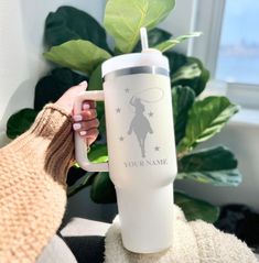 a person holding a white coffee mug with the name your name on it and a green plant in the background