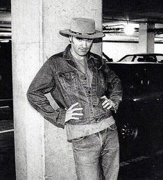 black and white photograph of man in hat leaning against wall with hands on hips while looking at camera