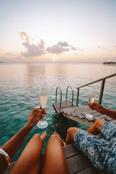 two people sitting on a dock holding wine glasses in front of the ocean at sunset