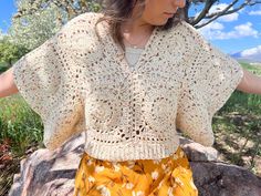 a woman standing in front of a tree wearing a yellow skirt and a white crochet sweater