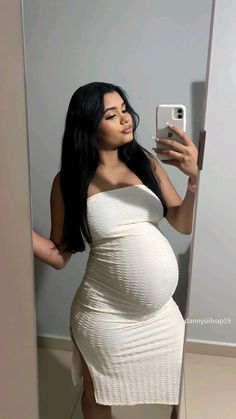 a pregnant woman taking a selfie in the mirror with her cell phone while wearing a white dress