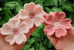 two crocheted flowers are held in front of some green leaves and bushes, one is pink and the other is light pink