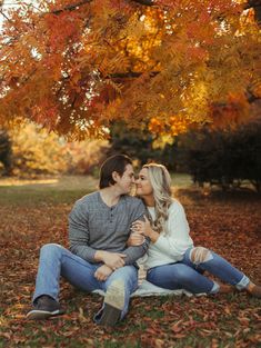 a man and woman sitting on the ground kissing under a tree with leaves all around them