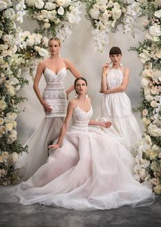 three women in white dresses posing for a photo with flowers on the wall behind them