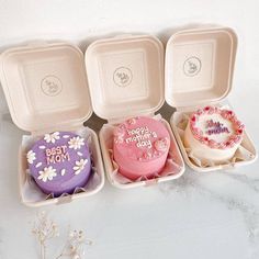 four decorated cakes in small trays on a table with white flowers and pink frosting