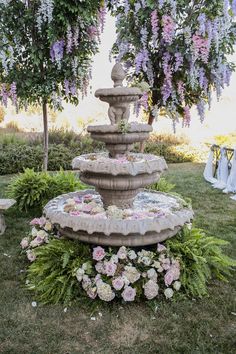 an outdoor fountain surrounded by flowers and greenery in front of a tree with purple wisters