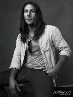 a man with long hair sitting on top of a wooden chair in front of a black and white photo