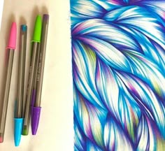 three colored pens are next to an art piece with feathers on it and another drawing is in the background