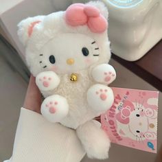 a white hello kitty stuffed animal with pink ears and paws holding a small card in it's hand