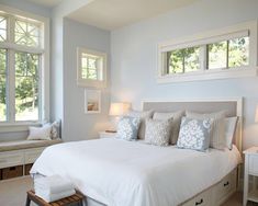 a white bed sitting in a bedroom next to two windows with lots of pillows on top of it