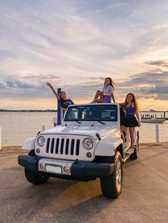 three girls standing on top of a white jeep near the water at sunset or dawn