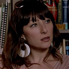 a woman with dark hair and large earrings looks at the camera while standing in front of bookshelves