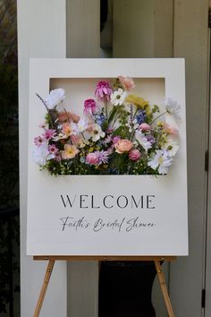 a welcome sign with flowers on it in front of a door and a wooden easel