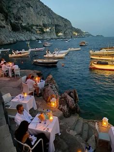 people are sitting at tables near the water with boats in the background and lit candles on them