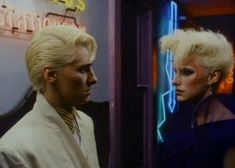 two people standing next to each other in front of a neon sign and the man has blonde hair