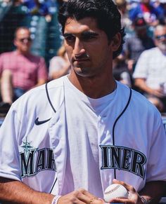 a man holding a baseball in his right hand and wearing a white shirt with the word mariners on it