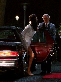 an older man standing next to a woman getting out of a red car at night