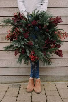 a woman holding a wreath with red berries and greenery on it in front of a wooden wall