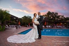 a bride and groom standing in front of a swimming pool at sunset with the sun setting behind them