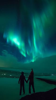 two people holding hands in front of an aurora bore over the ocean with green and blue lights