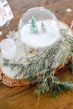 a wooden table topped with a glass bowl filled with snow and pine trees on top of it