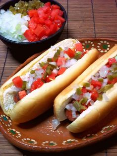 two hot dogs with toppings on a plate next to a bowl of tomatoes and lettuce