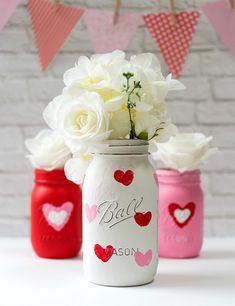 three painted mason jars filled with white and pink flowers on a table next to bunting flags