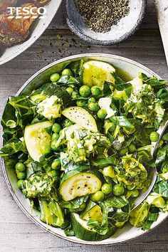 a white bowl filled with green vegetables next to other dishes and spoons on top of a wooden table