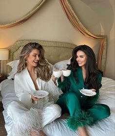 two beautiful women sitting on top of a bed holding cups and saucers in their hands