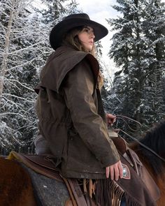 a woman wearing a hat riding on the back of a brown and black horse in snow covered forest