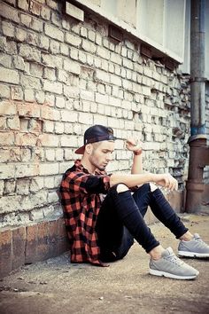 a man sitting on the ground next to a brick wall with his hands in his pockets