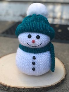 a small knitted snowman sitting on top of a wooden stump wearing a green hat and scarf