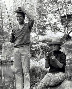 a man and woman sitting on rocks with fishing rods in their hands, while one holds a fish