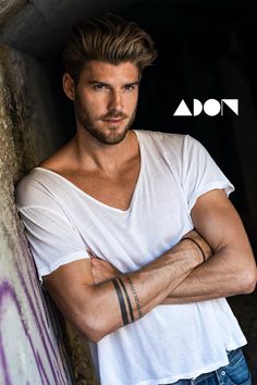 a man with his arms crossed leaning against a wall and looking at the camera while wearing a white t - shirt