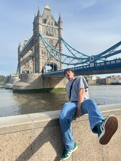 a man sitting on the edge of a stone wall in front of a suspension bridge