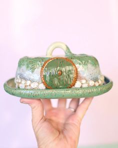 a hand holding a green ceramic dish with pearls and a ring on the top, in front of a pink wall