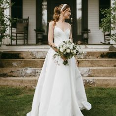 a woman standing in front of a house wearing a wedding dress and holding a bouquet